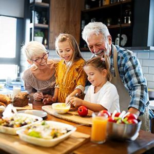Two smiling grandparents and two young girls prepare food together in a bright kitchen, surrounded by dishes and fresh vegetables. The group appears happy and engaged in a cooking activity.